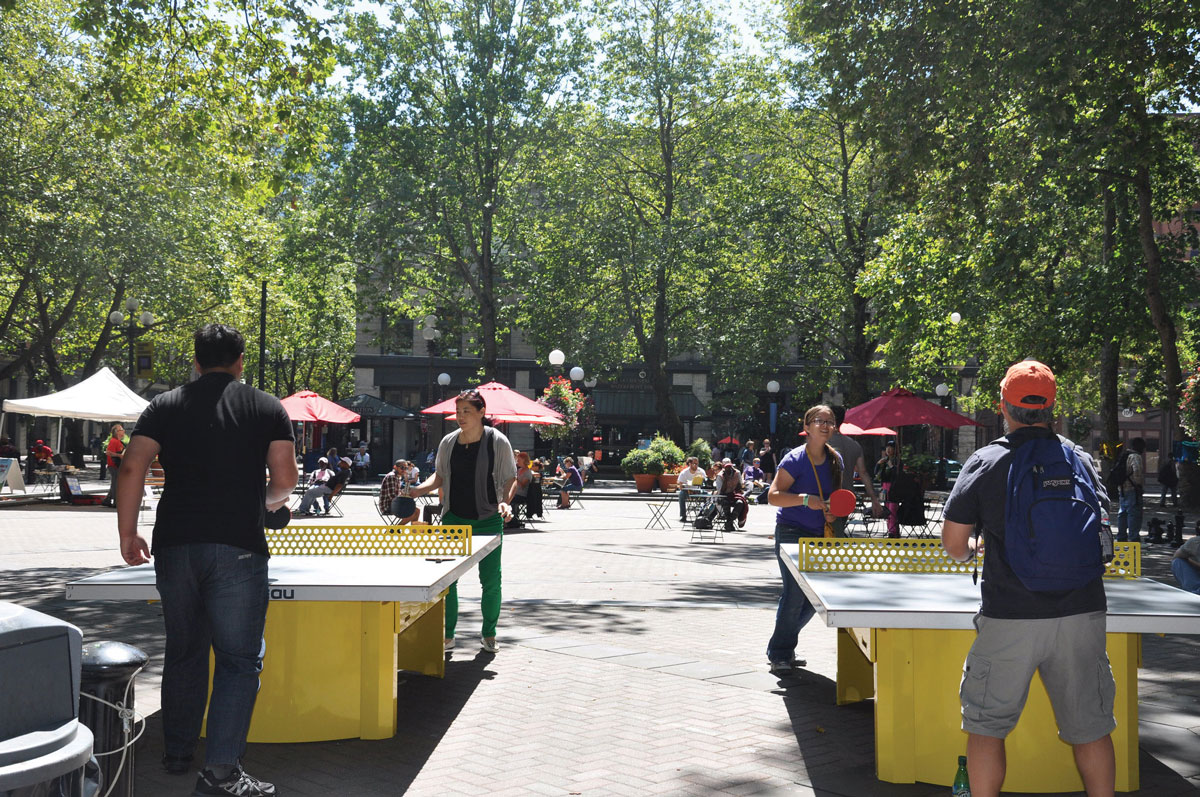 People play table tennis on yellow tables in a sunny outdoor plaza surrounded by trees, with others sitting at tables under red umbrellas in the background.