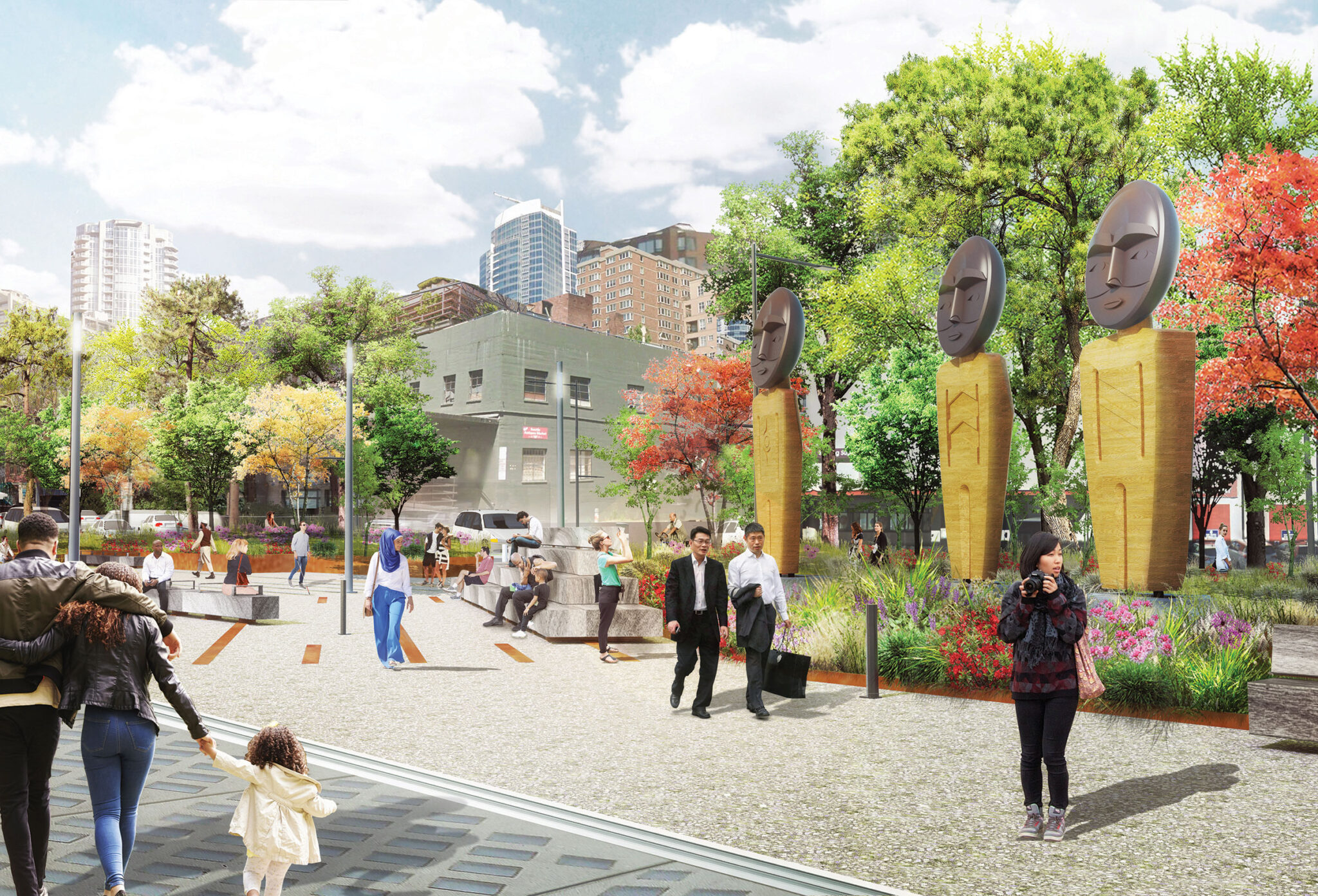 A lively city park with people walking, sitting, and taking photos. The park features three tall wooden sculptures with abstract face designs, colorful trees, and modern buildings in the background under a partly cloudy sky.