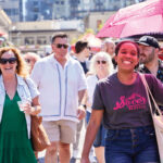 A group of people walk outdoors on a sunny day. A smiling woman in front holds a pink "Savor Seattle" umbrella, while others behind her wear sunglasses and summer clothes. Urban buildings and market stalls are in the background.