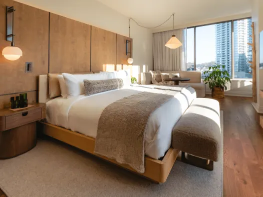 Modern hotel room with a large bed, neutral decor, wooden accents, a plush bench, and a sitting area near wide windows overlooking city buildings. Natural light fills the space, and a leafy plant sits by the window.