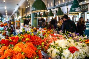 Bright bouquets of orange, yellow, and white flowers fill tables at an indoor market.