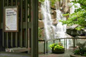 A sign for Waterfall Garden Park is posted on a gate beside a lush courtyard with a waterfall.