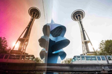 The Space Needle in Seattle is seen next to a mirrored building reflecting its image, with a monorail passing by in the foreground.