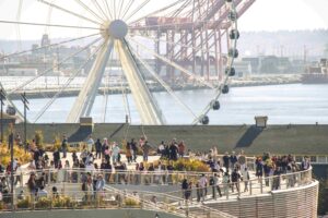 A large crowd of people walks along a waterfront promenade with a Ferris wheel and industrial structures in the background.
