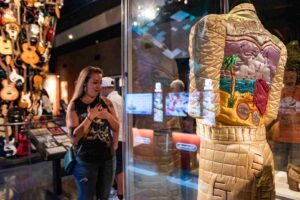 A woman stands in awe, looking at an embroidered jumpsuit displayed in a glass case at a museum, with guitars hanging on the wall in the background.