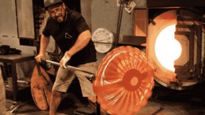 A man wearing glasses and a cap shapes molten glass on a long rod in a workshop, rotating it near a glowing furnace. 