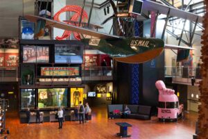 A museum interior featuring a suspended green U.S. Mail glider, people interacting below, a giant pink hand sculpture on a van.
