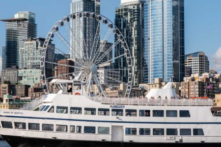 A white Argosy Cruises ferry sails on the water in front of the Seattle skyline, featuring modern high-rise buildings and the Seattle Great Wheel under a clear blue sky.