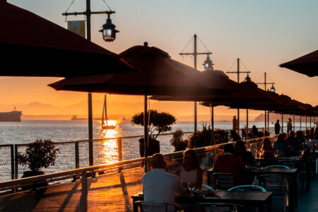 People dine at outdoor tables under large umbrellas on a waterfront patio at sunset. The sky glows orange, sailboats and ships are visible on the water, and distant mountains create a scenic backdrop.