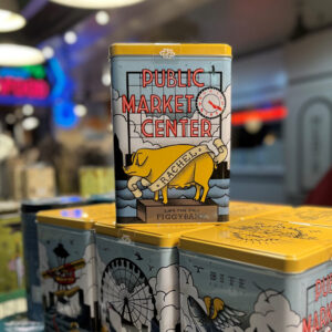 A rectangular tin piggy bank decorated with the Pike Place Market sign and a yellow pig labeled "Rachel" is displayed on a stack of similar tins in a souvenir shop. The background is colorful and blurred.