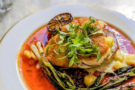 A plated dish featuring a seared fish fillet topped with fresh herbs, served with grilled green onions, diced potatoes, a charred lemon half, and a vibrant red sauce on a white plate.