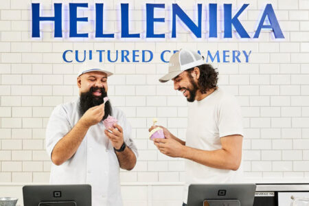 Two men stand smiling behind a counter at Hellenika Cultured Creamery, sharing a dessert. One wears a chef’s coat and hat, the other a t-shirt and baseball cap. Both seem to enjoy the moment in the bright, modern shop.
