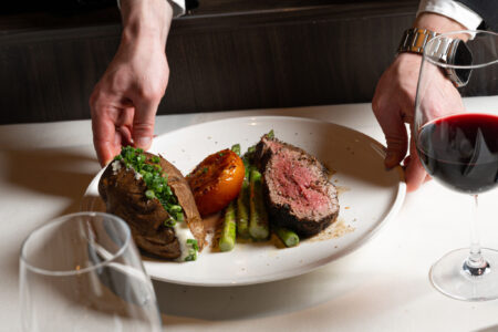 A person in formal attire serves a plate with a baked potato, grilled tomato, asparagus, and a slice of rare steak. A glass of red wine and an empty wine glass are visible on a white tablecloth.