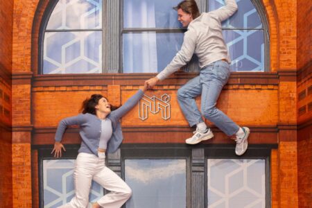 A woman and a man appear to hang from a large brick building facade, holding hands and smiling. The scene creates an illusion of climbing the wall, with both dressed casually in jeans and sweaters.
