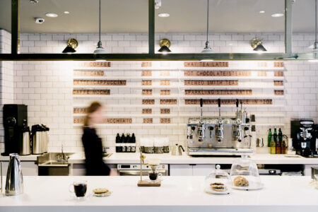 A modern coffee shop with white tiled walls, a wooden menu board, coffee machines, pastries under a glass dome, and a blurred person walking behind the counter.