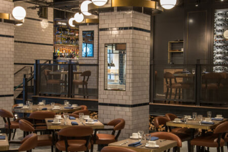 Modern restaurant interior with brown leather chairs, wooden tables set with glasses, plates, and napkins, white tiled columns, globe pendant lights, and a visible bar area in the background.