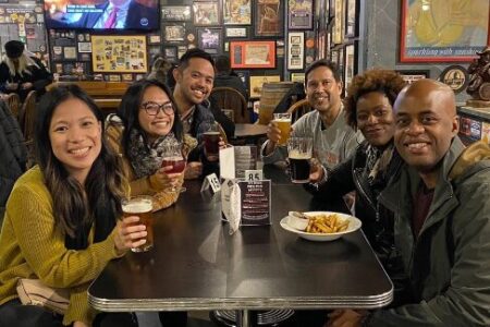 Six friends sit around a table at a restaurant, smiling and holding drinks. There are fries on the table, and the walls behind them are covered with framed photos and posters. A TV is on in the background.