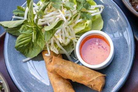 Two golden-brown spring rolls on a gray plate, served with fresh basil, lettuce, bean sprouts, cilantro, and a small dish of orange dipping sauce.