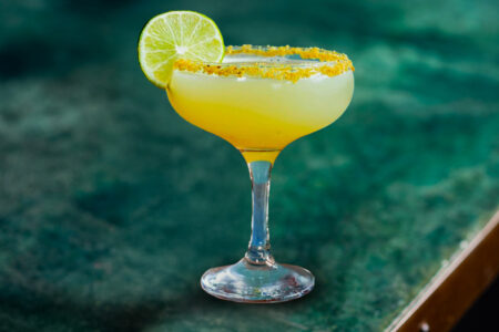A yellow cocktail in a coupe glass, garnished with a lime wheel and a spiced salt rim, sits on a green marbled surface.