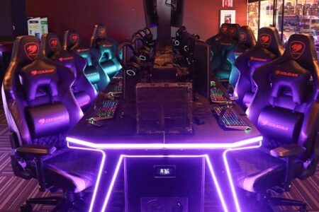 A row of high-end gaming chairs and RGB-lit desks arranged in a modern gaming lounge or esports center, with multiple monitors, keyboards, and gaming accessories visible. The room has a futuristic ambiance.