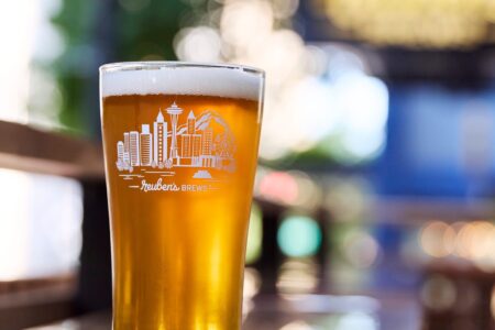 A pint glass of amber beer with a white foam head sits on a wooden table. The glass features a white illustration of a city skyline, trees, and the text “Zeuben’s Brews.” The background is softly blurred.