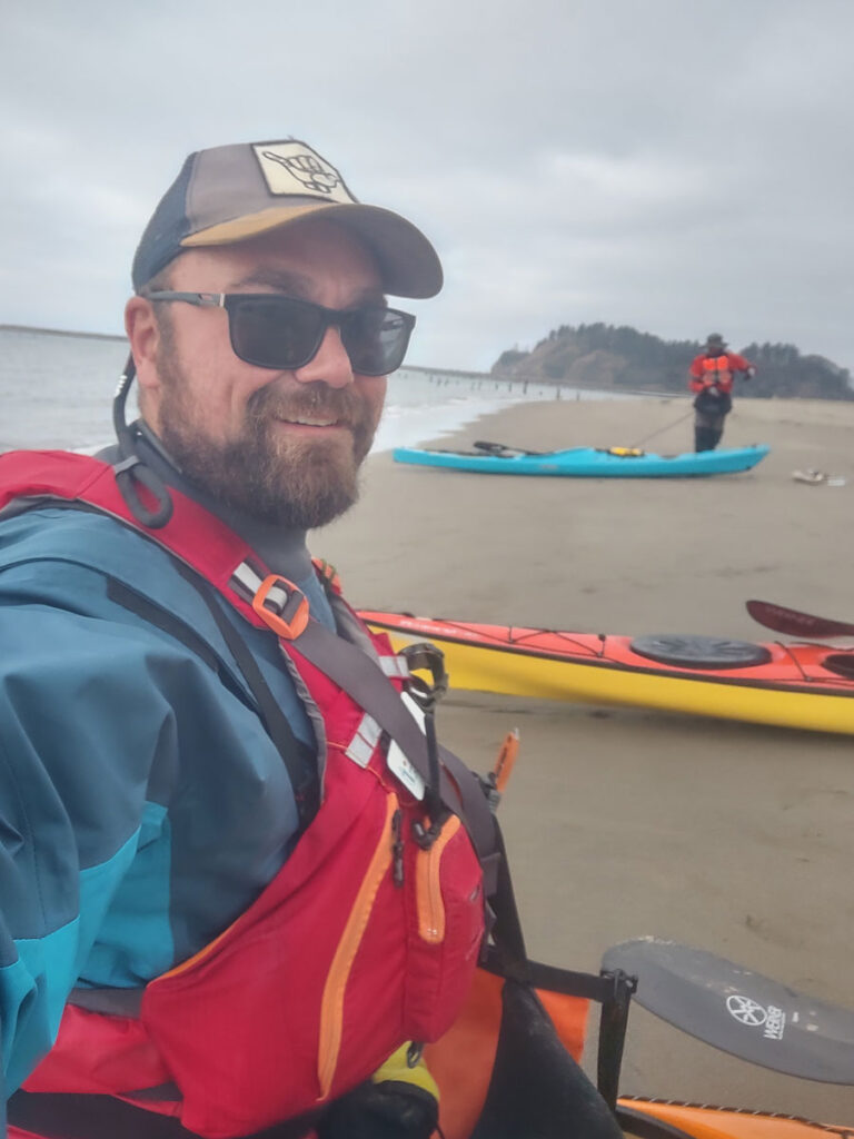 A man in sunglasses and a life vest takes a selfie on a sandy beach with kayaks and another person in the background under a cloudy sky.