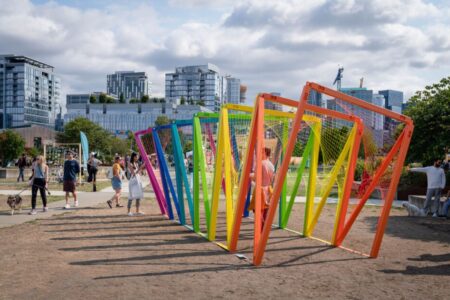 People walk and interact around a colorful outdoor art installation made of rainbow-colored frames in a city park, with modern buildings and trees in the background on a partly cloudy day.