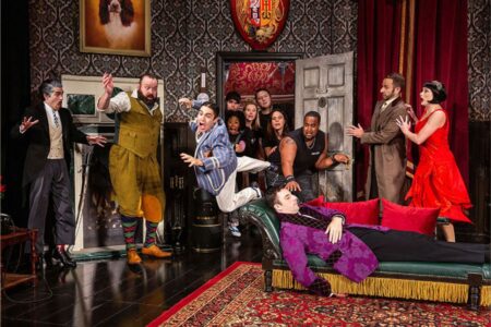 A lively stage scene shows actors in colorful costumes reacting dramatically as a man in pajamas appears to trip and fall over a fainted person on a couch, while others watch in shock or try to help. The setting is an ornate room.