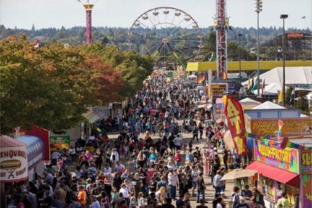 A large crowd walks through a busy fairground lined with colorful food stalls and games. In the background, a ferris wheel and rides are visible among trees under a clear sky.