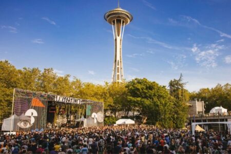 A large outdoor crowd attends a music festival with a colorful stage beneath the Space Needle in Seattle, surrounded by trees and blue sky.