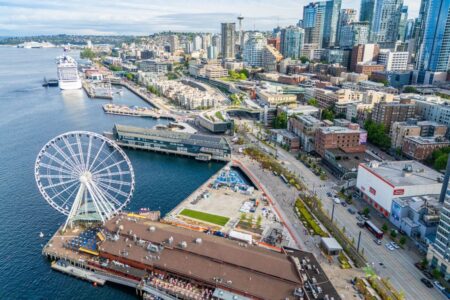 Aerial view of Seattle’s waterfront featuring the Seattle Great Wheel, piers, cruise ships, and downtown skyscrapers with Elliot Bay and distant hills in the background.