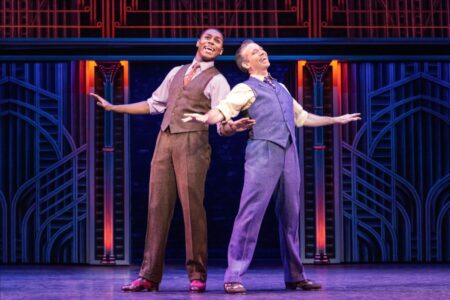 Two actors in vintage suits stand back-to-back on stage, smiling with arms outstretched. The colorful, art deco-style set behind them is illuminated with vibrant lights.