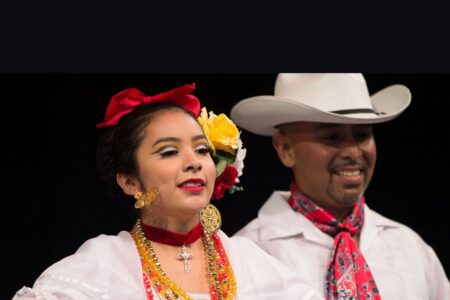 A woman and man in traditional Mexican attire smile while performing. The woman wears a white blouse, elaborate jewelry, a red bow, and yellow flowers in her hair. The man wears a white cowboy hat, white shirt, and red scarf.