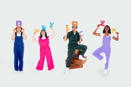 Four people in colorful jumpsuits hold up illustrated letters above their heads, each with eyes and faces. They stand or pose against a plain white background with one person sitting on stacked suitcases.