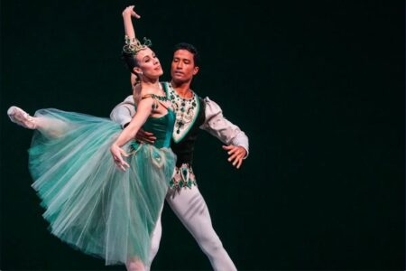 A male and female ballet dancer perform on stage. The female dancer wears a green tutu and jeweled headpiece, gracefully extending one arm, while the male dancer supports her, wearing a matching green and white costume.