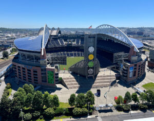 Aerial view of Lumen Field, a large stadium with a distinctive curved roof, open field, and empty seats, located in Seattle on a clear sunny day—a landmark for Seattle sports—surrounded by parking lots and greenery.