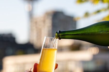 A hand holds a champagne glass as sparkling wine is poured outdoors, with a blurred cityscape and the Space Needle visible in the background on a sunny day.