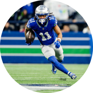A football player in a blue Detroit Lions uniform with the number 11 runs on the field holding a football, appearing to dodge an opponent during a game.