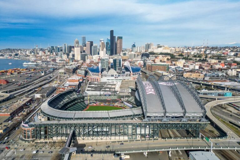 Aerial view of a baseball stadium with a partially open retractable roof, set against the backdrop of a city skyline with tall buildings and blue sky.