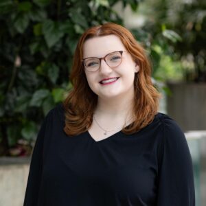 A woman with long red hair and glasses, wearing a black top, smiles while standing outdoors in front of green leafy plants.