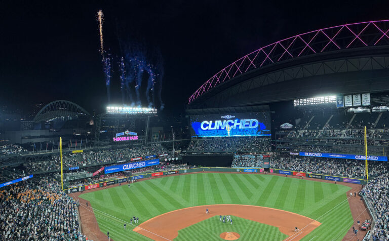 A packed Seattle sports baseball stadium at night bursts with fireworks. The scoreboard shows “CLINCHED,” and the well-lit field is surrounded by cheering fans. The partially open roof is aglow with purple lights, capturing the city’s vibrant energy.