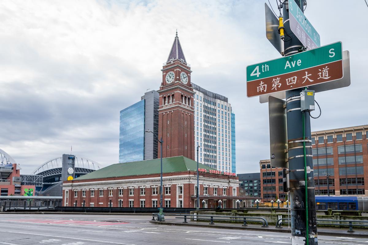 A street sign for 4th Ave S in English and Chinese is in the foreground, with Seattle’s King Street Station and its tall clock tower visible behind it under a cloudy sky.