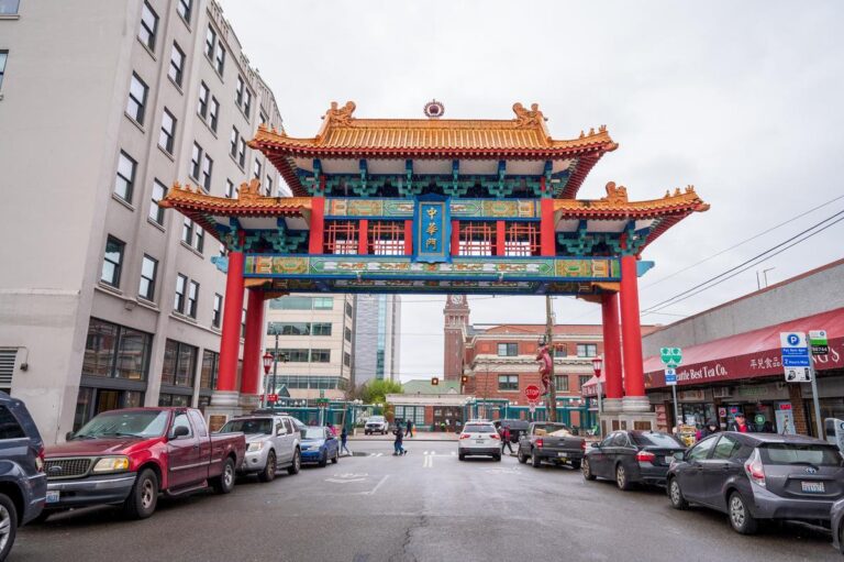A colorful traditional Chinese gate stands over a street lined with parked cars and surrounded by buildings on a cloudy day. The gate has ornate red columns, blue and gold accents, and a tiled roof.