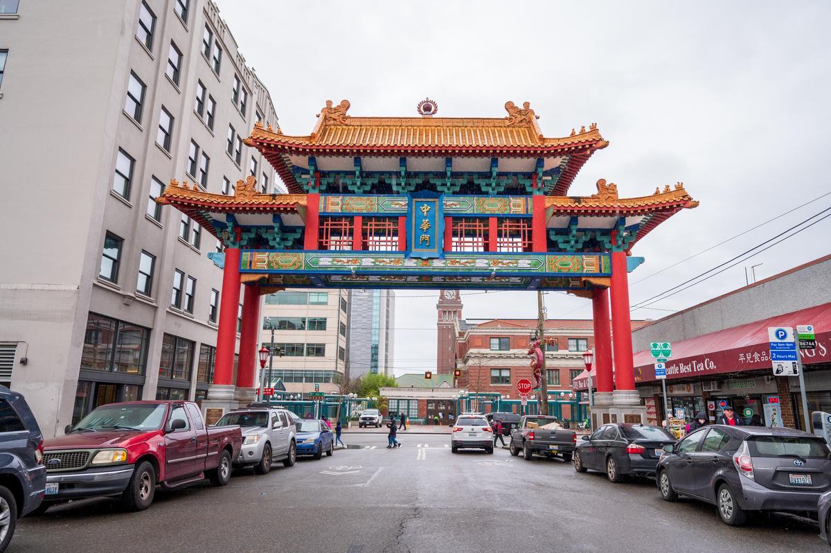 A colorful traditional Chinese gate stands over a street lined with parked cars and surrounded by buildings on a cloudy day. The gate has ornate red columns, blue and gold accents, and a tiled roof.
