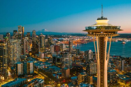 Aerial view of Seattle at dusk, featuring the illuminated Space Needle in the foreground, with city buildings, waterfront, and distant mountains under a clear sky.