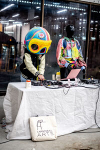 A person wearing a colorful helmet and varsity jacket mixes music on DJ equipment at a table, beside a mannequin dressed in bright, layered clothing. A "Cannonball Arts" tote bag hangs from the table.