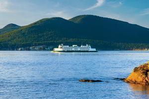 A white ferry boat sails across calm blue water with forested hills and mountains in the background under a clear sky. Rocky shoreline is visible in the foreground.