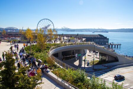People walk along a waterfront park with a spiral ramp, greenery, and views of a Ferris wheel and ships on a sunny day, as Seattle celebrates Indigenous Peoples' Day and Native American Heritage Month by the water.