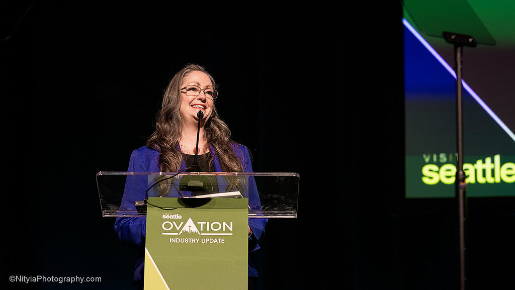A woman with long hair and glasses stands at a podium, smiling as she gives a speech during the Seattle Ovation Industry Update event. The background features a screen with colorful text and graphics.