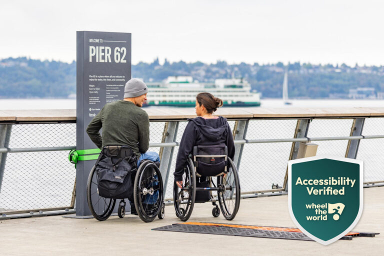 Two people in wheelchairs talk by a sign for Pier 62 on a waterfront boardwalk. A ferry passes on the water in the background. An “Accessibility Verified” badge is shown in the bottom right corner.